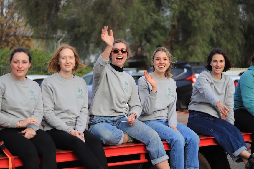 Five women sitting on the tray of a ute smile and wave.