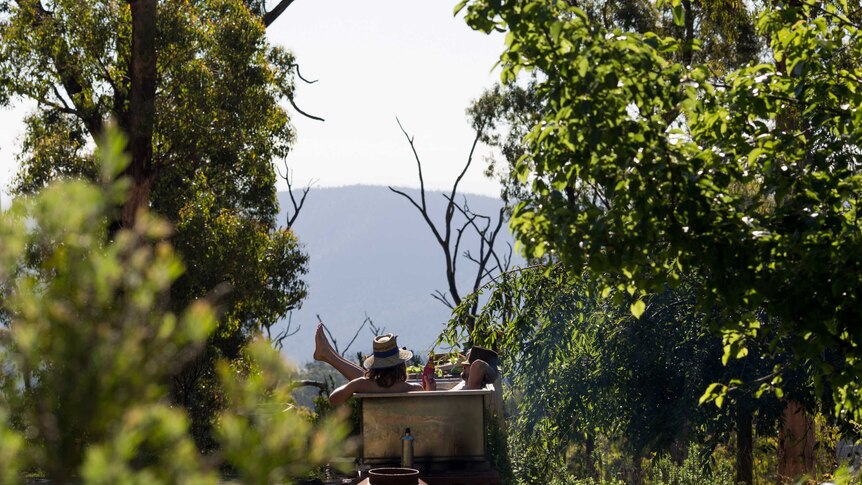 Two men with broad brim hats on their heads and beers in hand lounge in a bathtub surrounded by bush with mountains beyond.