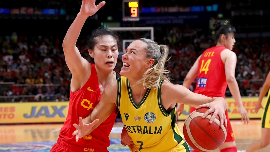 Tess Madgen grimaces and holds a basketball in one hand as a player in a red singlet holds her hand up next to her