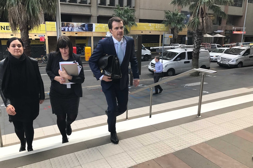 Jonathon Hunyor, dressed in a suit and holding a black bag under his arm, walks up a staircase accompanied by two women