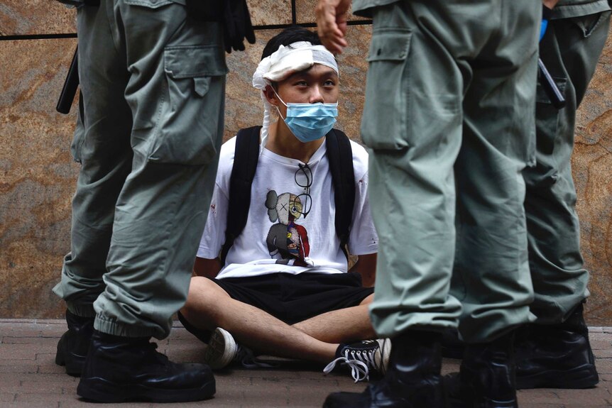 A man sit cross legged on the ground with a bandage on his head and a face mask is surrounded by soldiers' legs