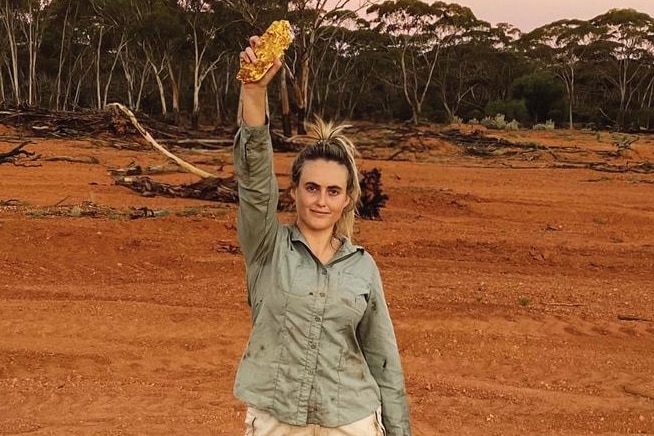 Tyler Mahoney, wearing kakhi shorts and shirt, and outdoor boots, stands in red dirt holding a large chunk of gold in her hand.
