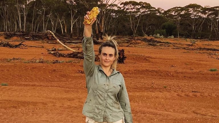 Young woman wearing kakhi shorts and shirt, and outdoor boots, stands in red dirt holding a large chunk of gold in her hand.