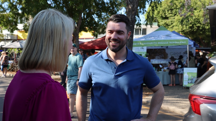 A woman and man speaking on the outskirts of an outdoor market, on a sunny day.