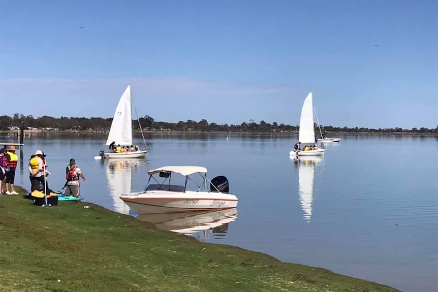 Girls wearing life jackets stand on the river, 3 boats with groups of people sail. The water and sky is bright blue.