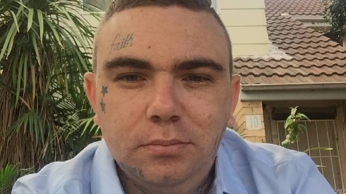 A man with short hair and a wry expression takes a selfie. He has the word "faith" tattooed on his head.