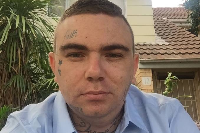 A man with short hair and a wry expression takes a selfie. He has the word "faith" tattooed on his head.