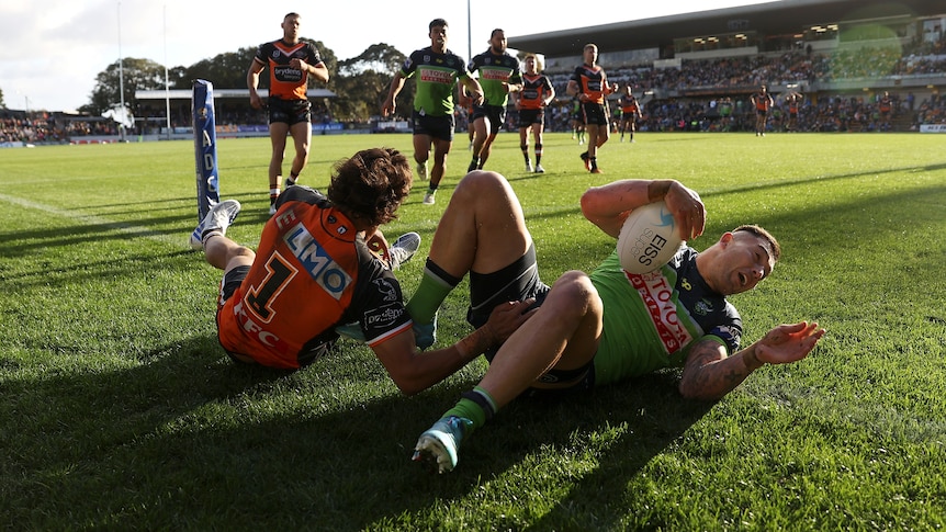 A Canberra Raiders NRL player lies on the ground holding the ball in the in-goal after a try, with a defender next to him.