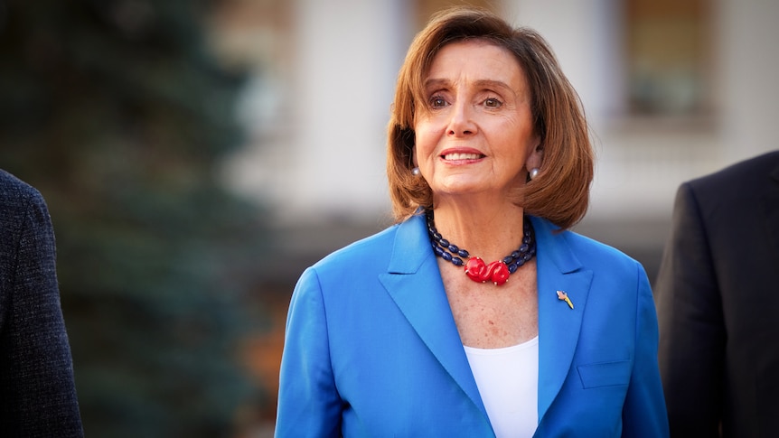 Nancy Pelosi in a statement black and red necklace and a bright blue suit with a slight smile on her face 