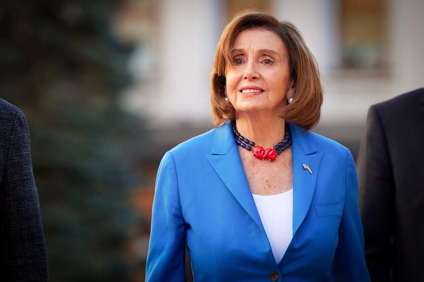Nancy Pelosi in a statement black and red necklace and a bright blue suit with a slight smile on her face 