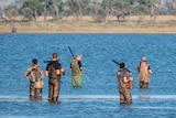 Five men, three holding guns, stand in water in camouflage clothing. Trees are in the background.