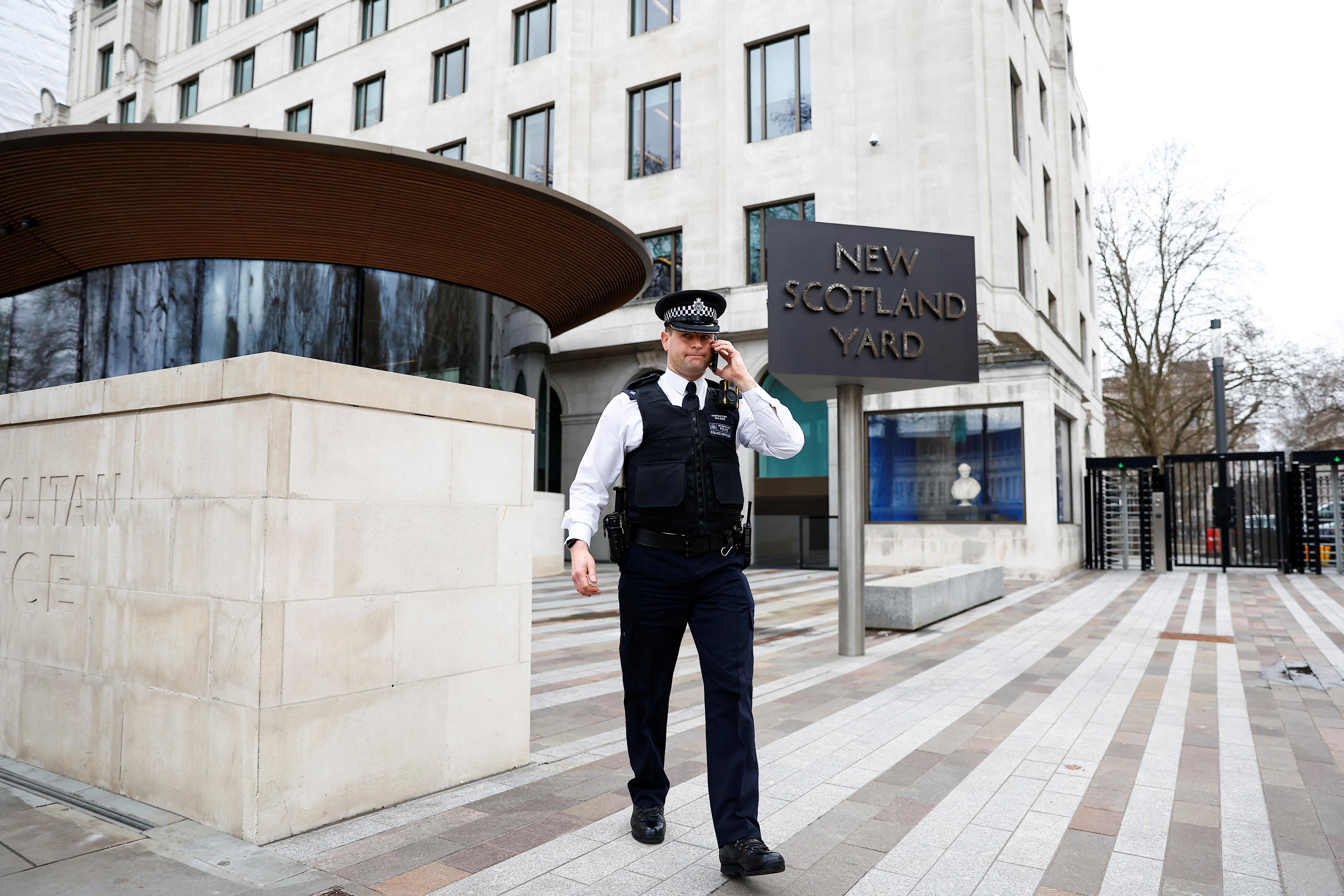 A British policeman in black and white clothing speaking on a mobile while walking next to an outdoor New Scotland Yard sign.