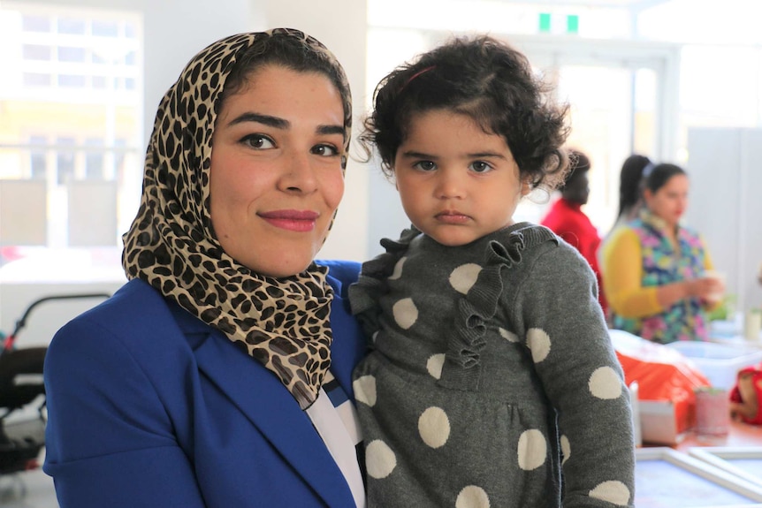 A woman wearing a leopard print headscarf and blue top smiles for a photo holding a young girl with dark hair and a grey top.