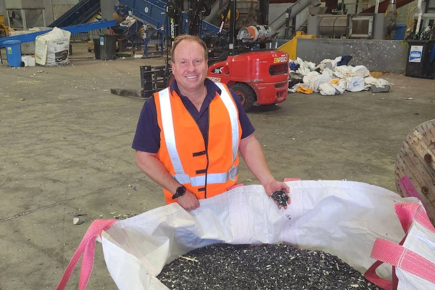 a man in a high-viz vest in an industrial shed standing next to a huge pile of recycled plastic