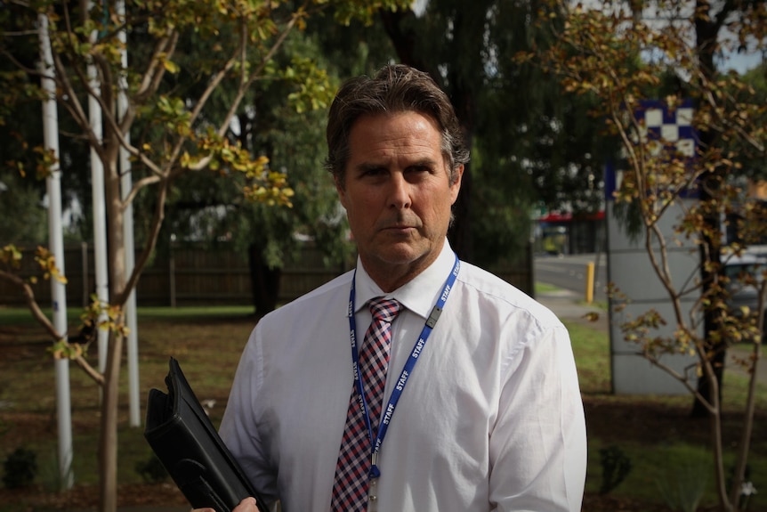A male detective in a shirt, tie and lanyard stands outside with a folder in his hand.