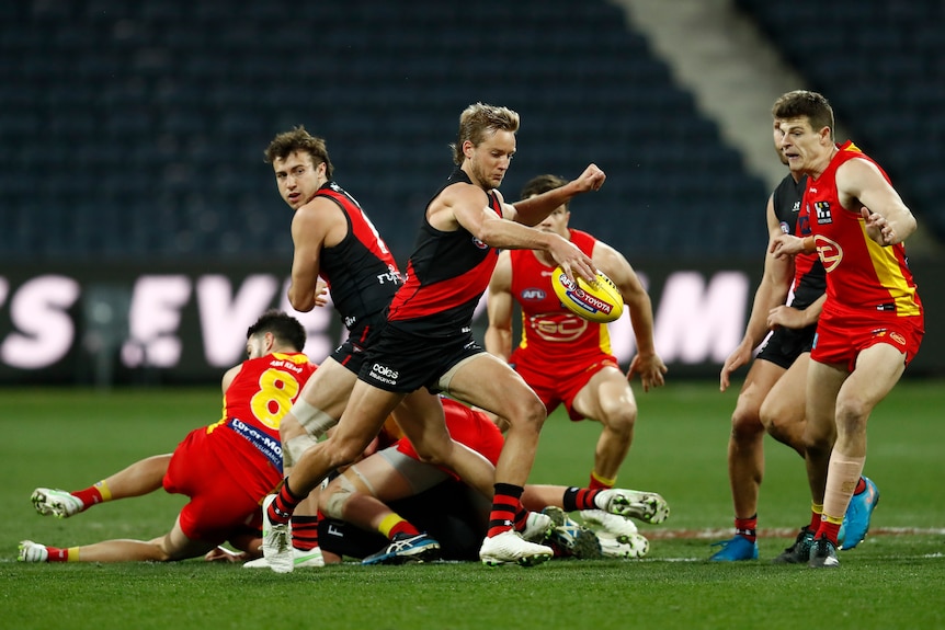 Un milieu de terrain d'Essendon regarde le ballon alors qu'il le frappe sur le terrain, tandis qu'un groupe d'autres joueurs est allongé sur le sol derrière lui.