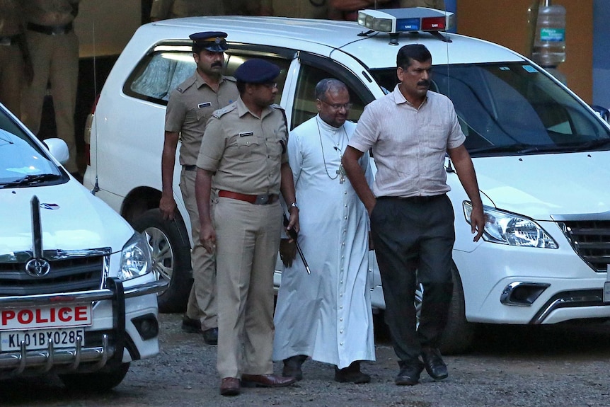 The Bishop is wearing a long white robe and walking, flanked by two police officers and another man.