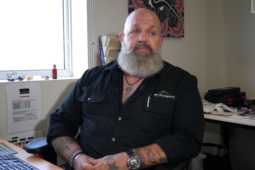 A man with a beard and tattoos sits at his desk in his office at a mining company.