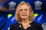 Queensland Chief Health Officer Jeannette Young wears glasses in front of a microphone, behind her is the Qld government logo.