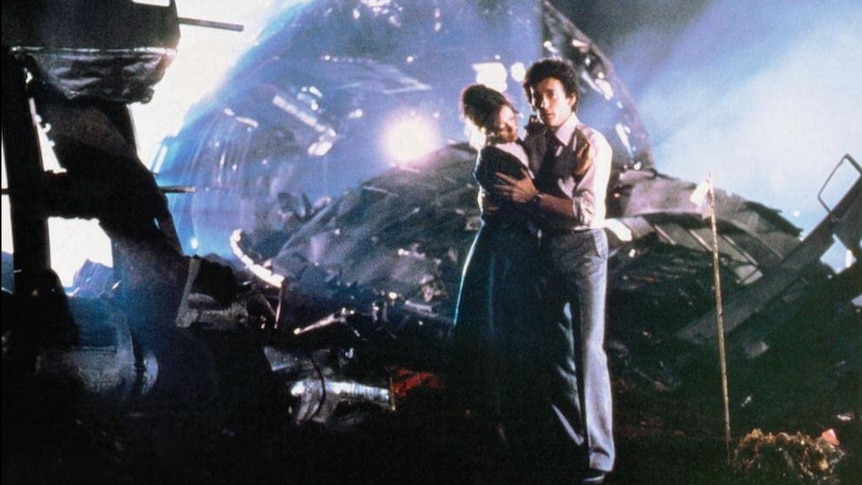 A man and woman hold each other in their arms with a plane crash wreckage in teh background