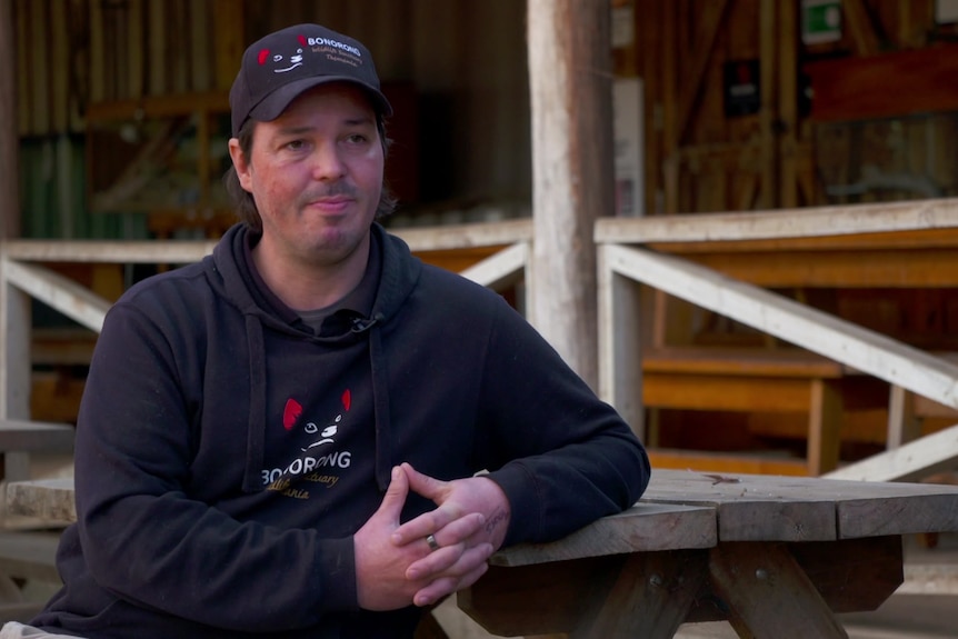 A man leans on a wooden post while talking to a reporter who is off-camera