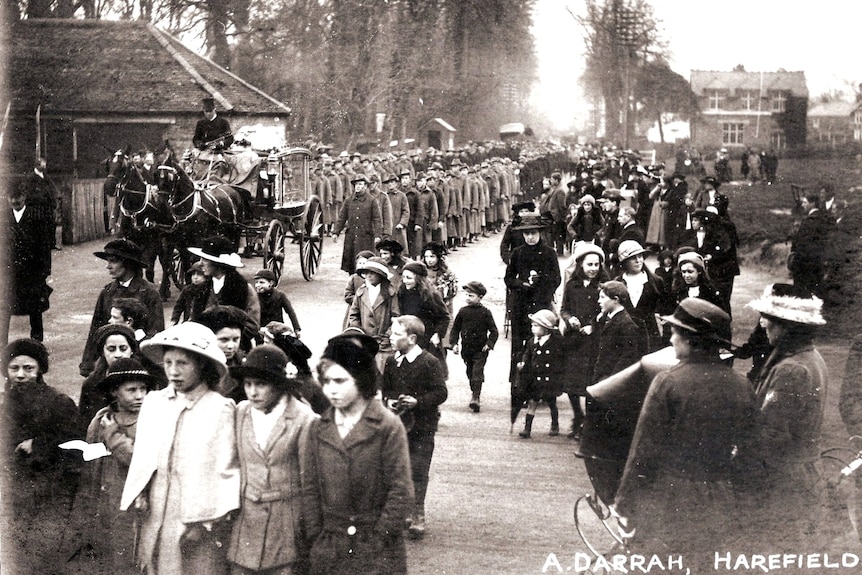 Men, women and children in winter clothing line a wide street, walking in the same direction.