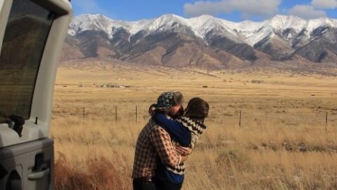 Jared Melrose and Ash embrace by their van with fields and snowcapped mountains in the background