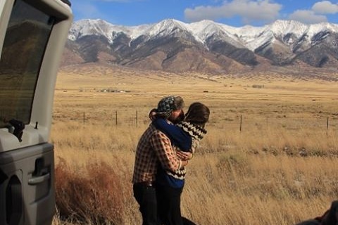 Jared Melrose and Ash embrace by their van with fields and snowcapped mountains in the background