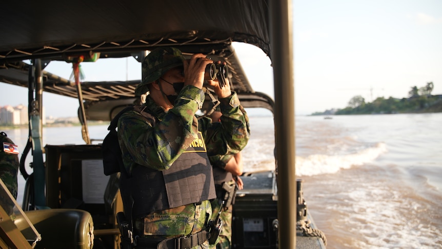 A man in camouflage gear stands on a boat, looking through binoculars