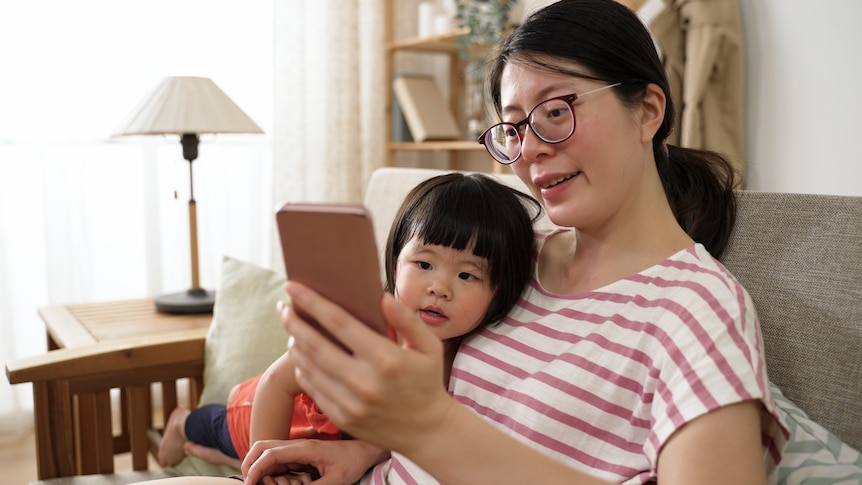 A woman sits on a lounge with a child, both of them looking at a smartphone in her hand.