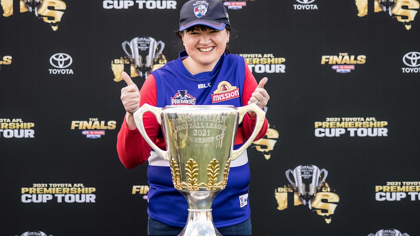 A woman wearing her AFL team's colours standing behind a trophy.  