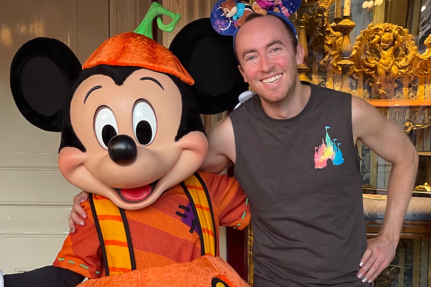 Joel Callen wearing a Disneyland tshirt and Disney ears puts his arm around someone in a Mickey Mouse jack-o-lantern costume.