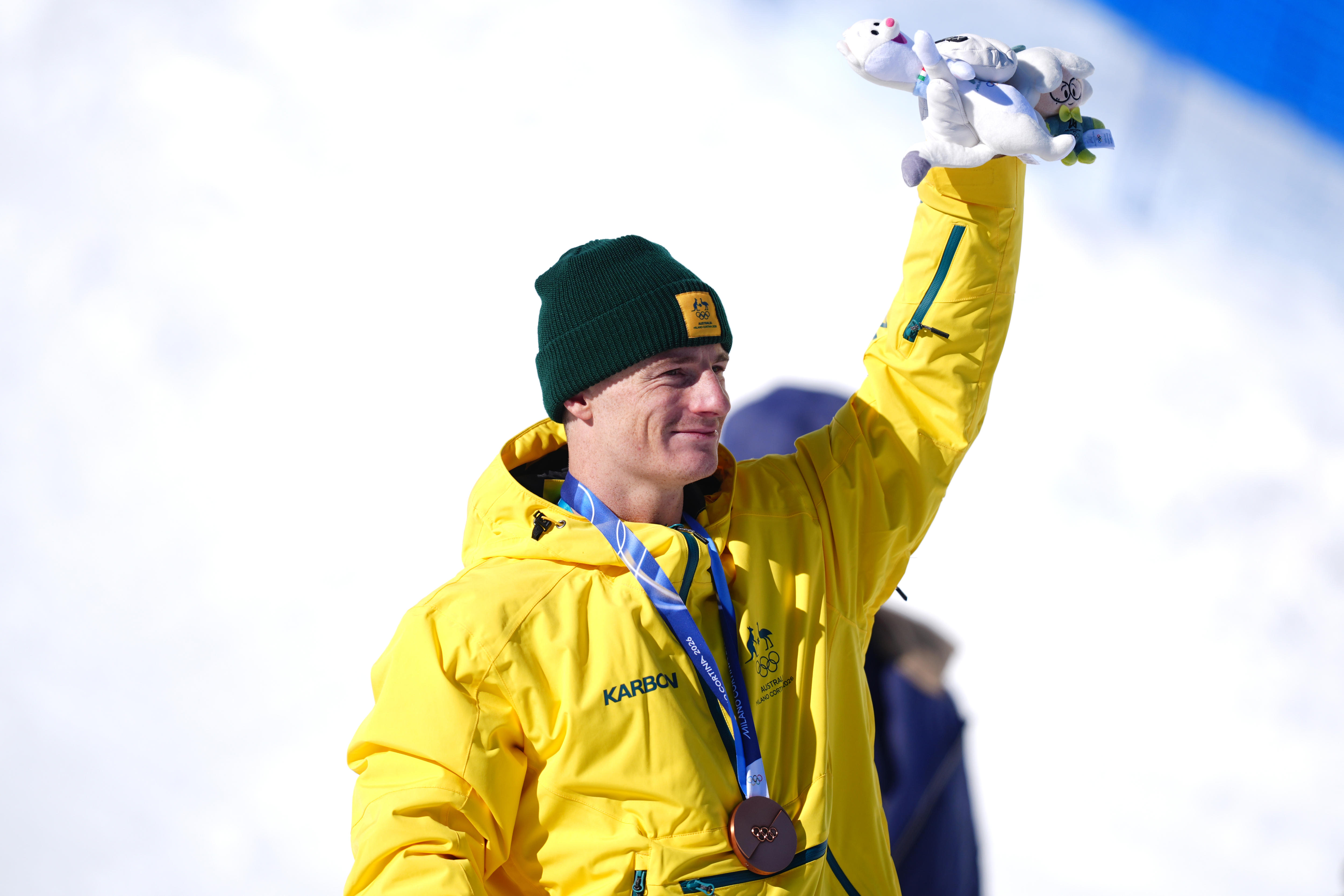 A man in a beanie and a parka with a medal around his neck holds up a plush toy while standing in a snowy area.