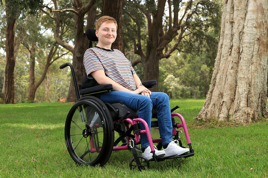 A young woman sits in her wheelchair. She wears a striped top, blue jeans and white runners and smiles. 