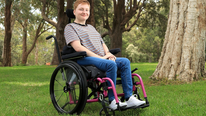 A young woman sits in her wheelchair. She wears a striped top, blue jeans and white runners and smiles. 