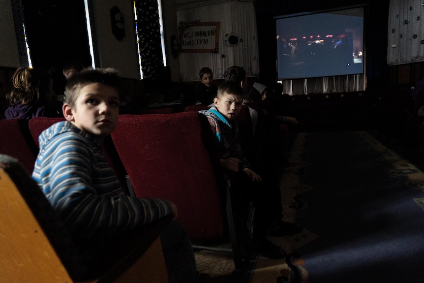 Two kids look over their shoulder as a movie plays in the background behind them.