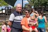 A couple holds a baby with other people in the background at a playground