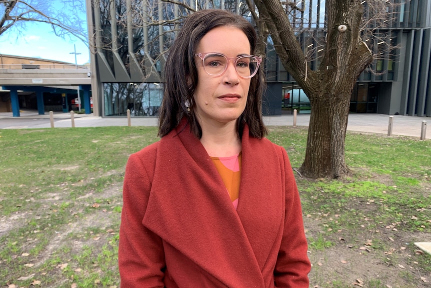 A woman stands in a park wearing pink glasses,earrings and a rust coat. Ausnew Home Care, NDIS registered provider, My Aged Care
