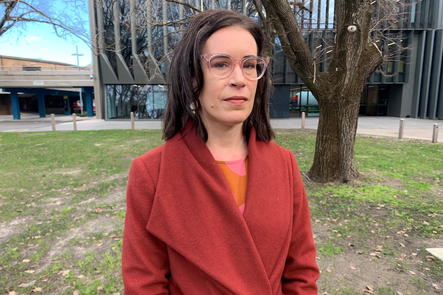 A woman stands in a park wearing pink glasses,earrings and a rust coat.