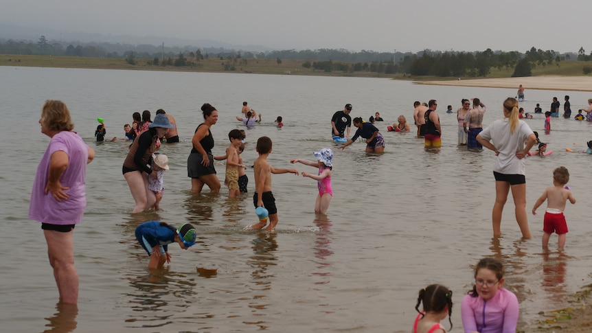 People stand in the shallows near the shore with a grey sky. 