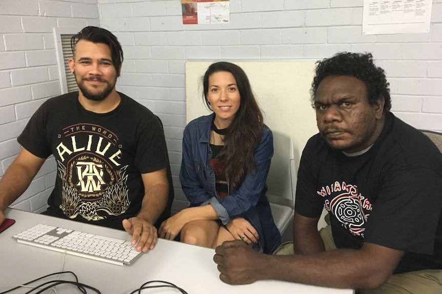 The three sit sit behind a desk, in front of a grey wall