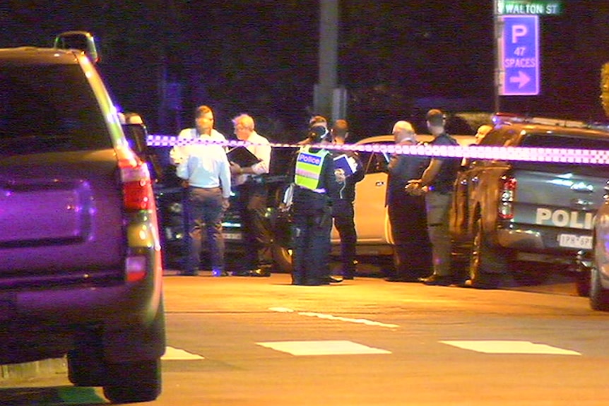 Uniformed police and detectives stand behind a police tape at night.