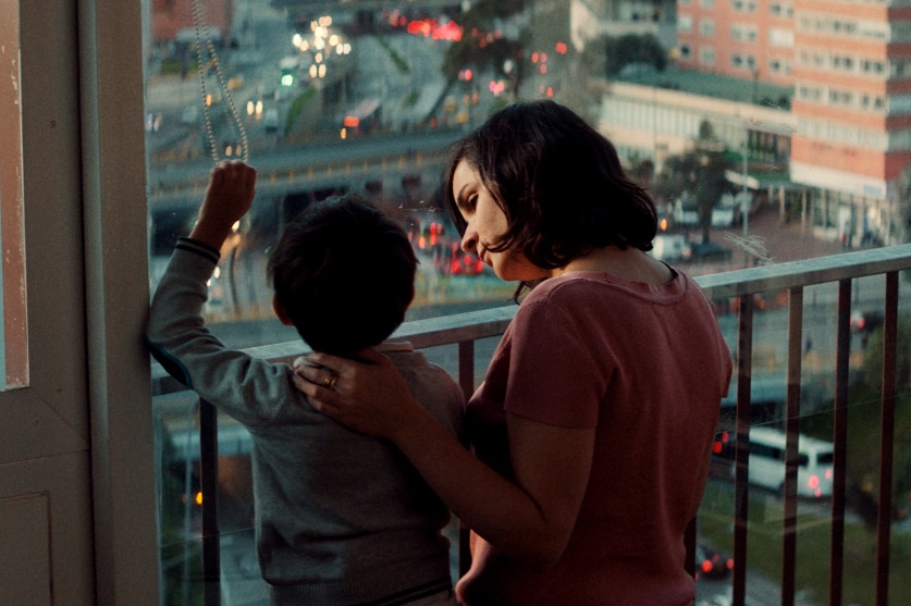 A woman stands and looks down to a young male child beside her, her hand supporting his neck as he looks out at dusk cityscape.