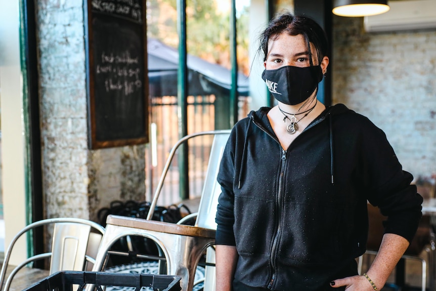 A young woman wearing a black mask and black zip jacket stands in an empty cafe.