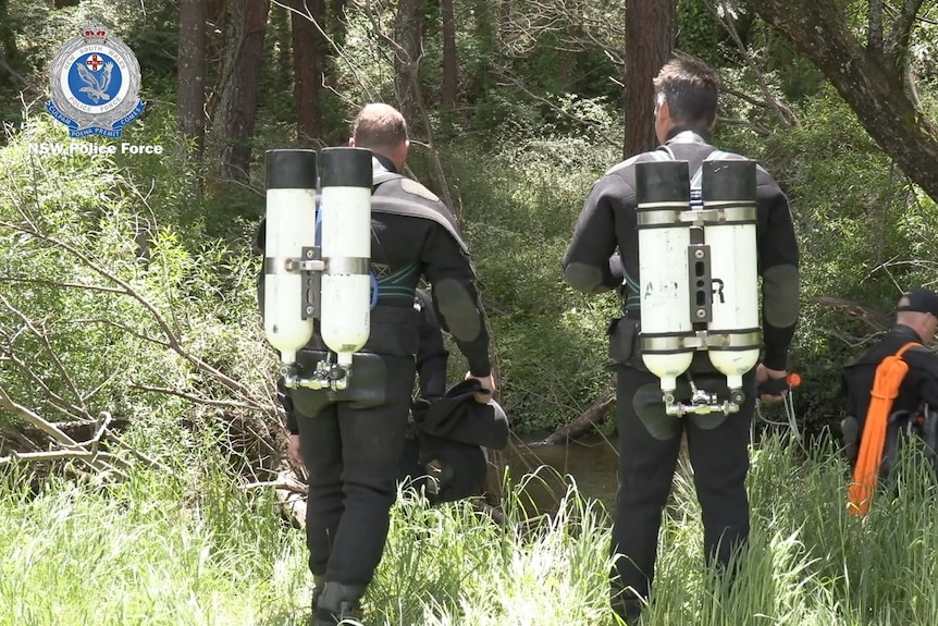 Two men wearing swim suits wear white oxygen tanks on their back and look on towards a creek.