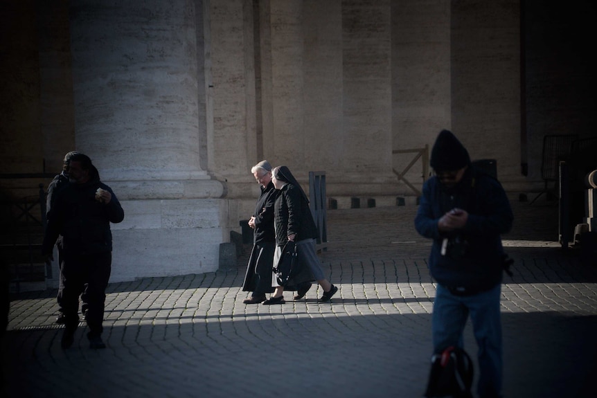 Two nuns chatting, viewed from a distance with large stone pillars in the background.