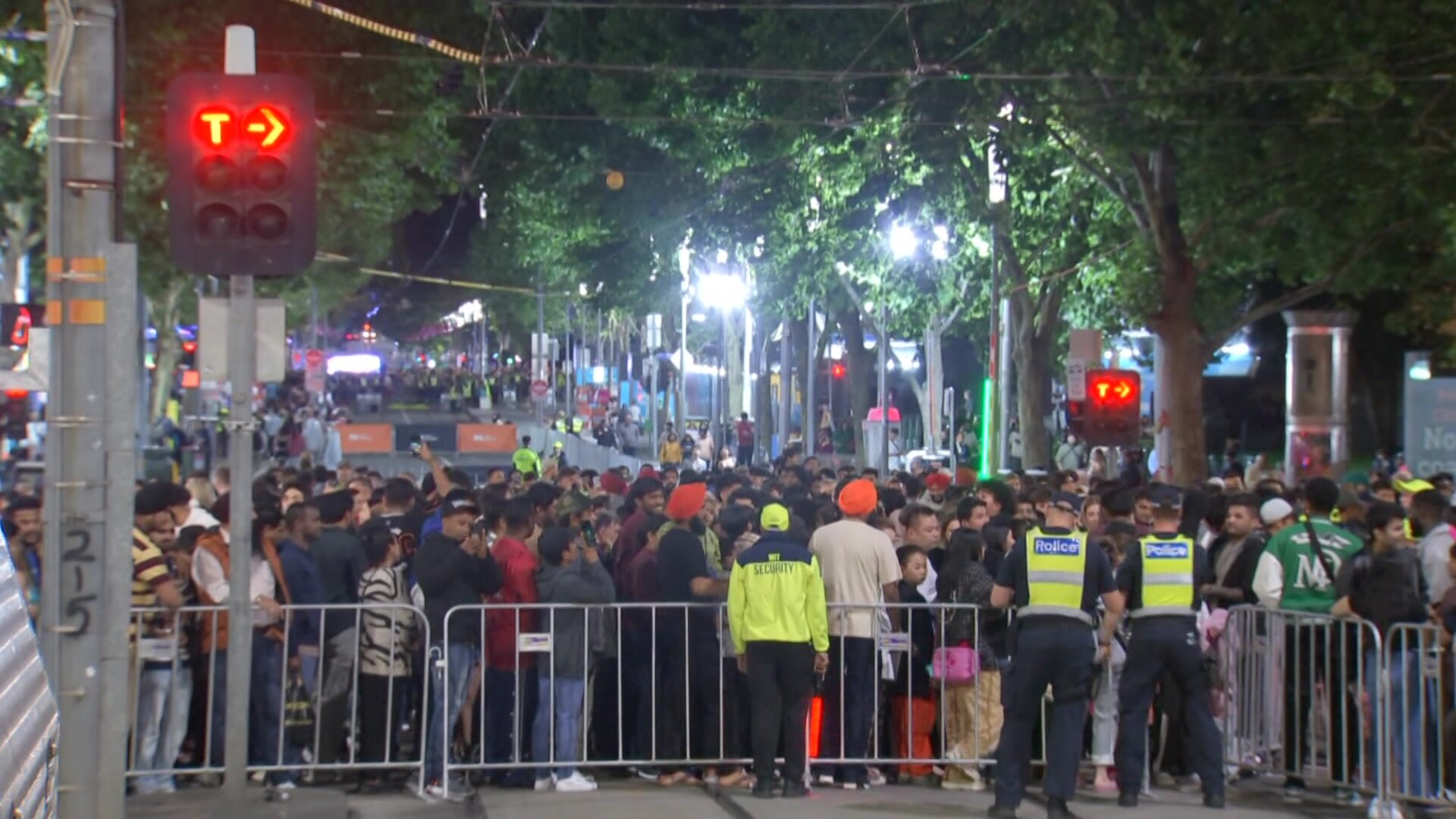 Policiais com coletes amarelos de alta visibilidade ficam diante de uma grande multidão atrás de uma cerca temporária em uma rua da cidade à noite.