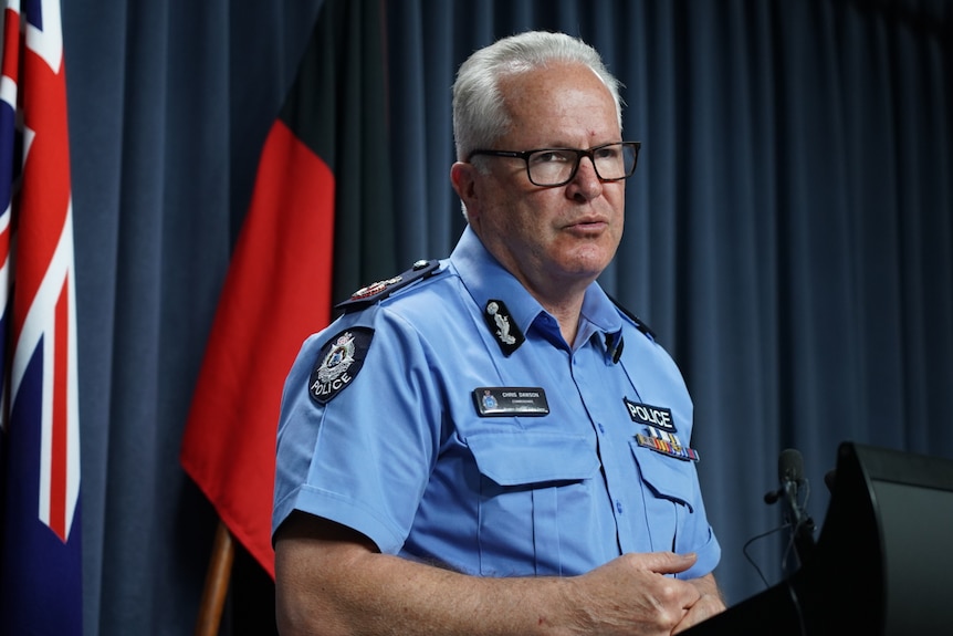 A man wearing glasses and a police uniform stands in front of a flag at a press conference