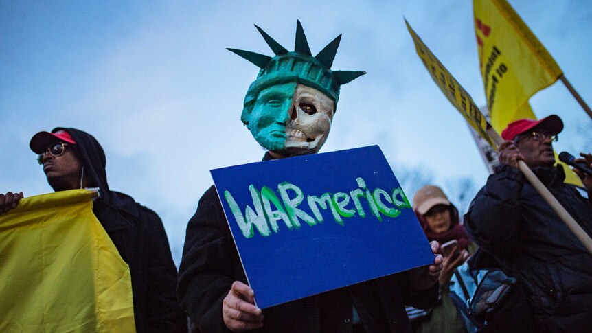 A protester holds a sign reading "Warmerica" and wears a mask that is half the statue of liberty and half a skull.