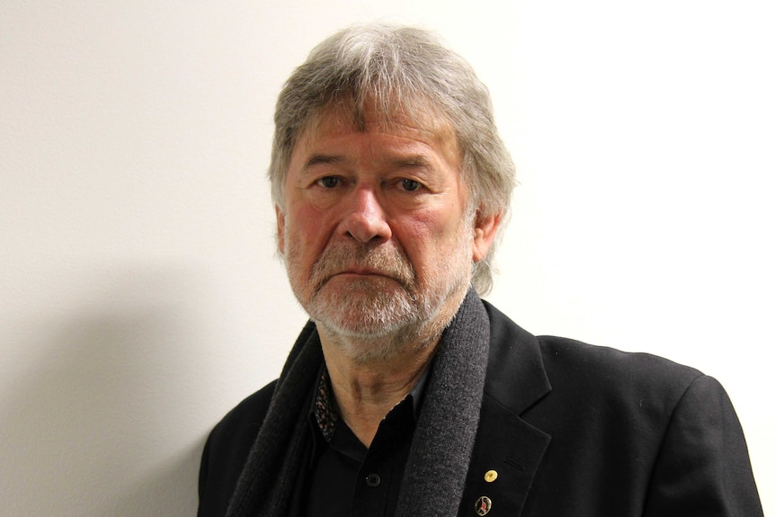 Older man with grey hair and short beard looks glumly at the camera.